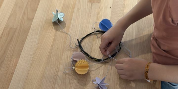 Man sieht kinderhände die einen großen Ring basteln an dem selbstgebastelte aus alten Papier hergestellte Dekorationen hängen.