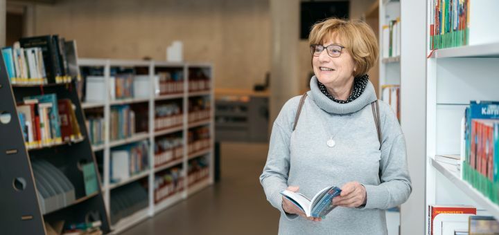 Ältere Frau steht mit einem Reiseführer in der Hand in der Bibliothek